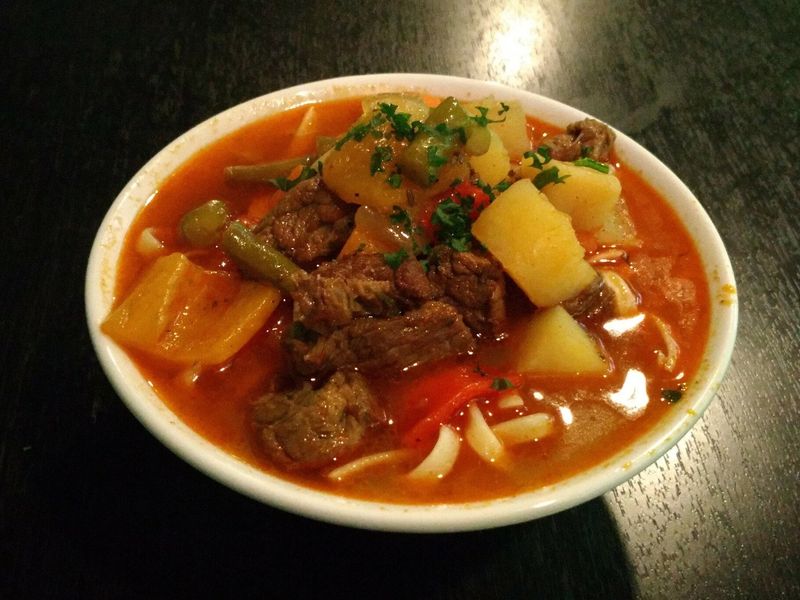 Лагман в белой миске — кусочки говядины, картофель, перец и стручковая фасоль в красном бульоне с лапшой и петрушкой