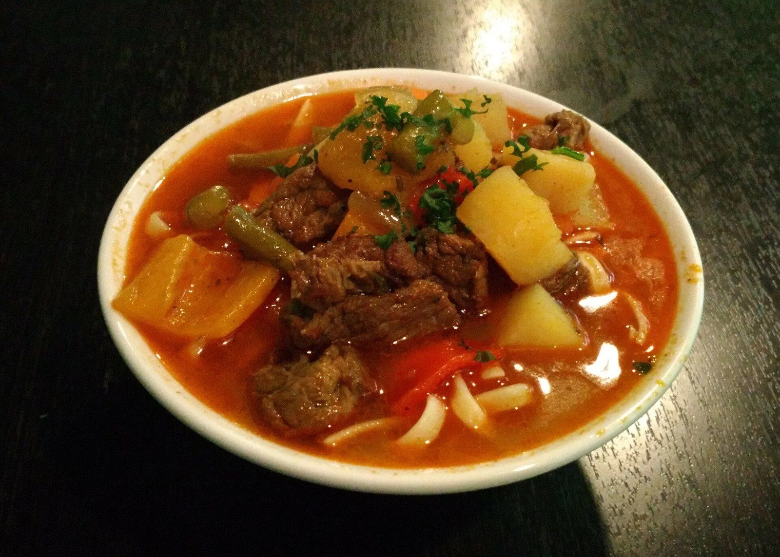 Лагман в белой миске — кусочки говядины, картофель, перец и стручковая фасоль в красном бульоне с лапшой и петрушкой